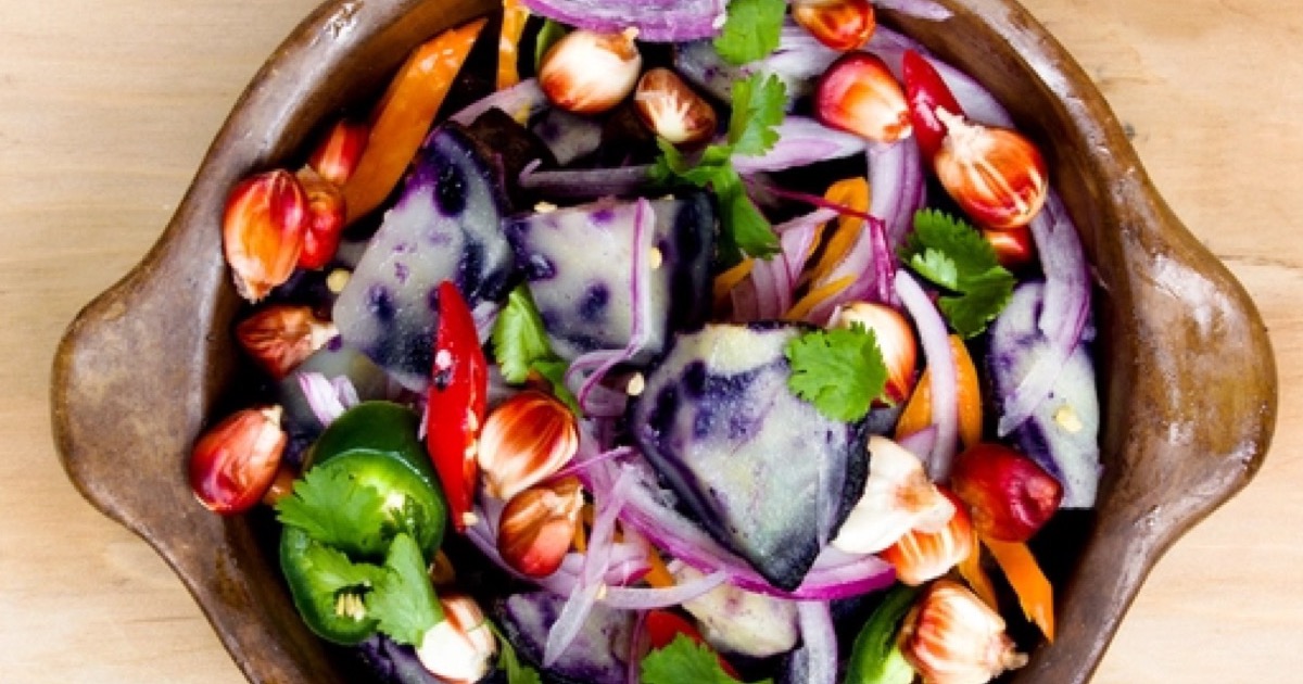 An overhead view of a wooden bowl of purple potato salad with red onion, peppers and cilantro.
