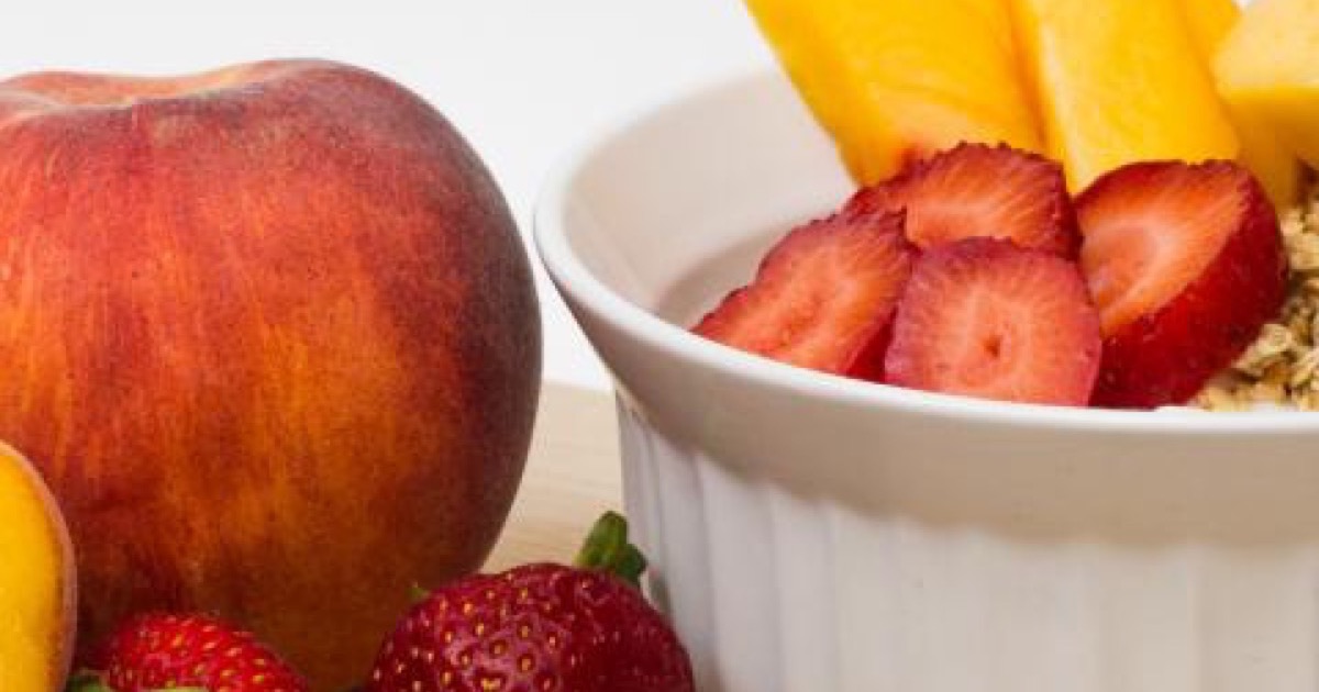 A white ramekin of sliced strawberries and mango beside a whole peach and fresh strawberries.