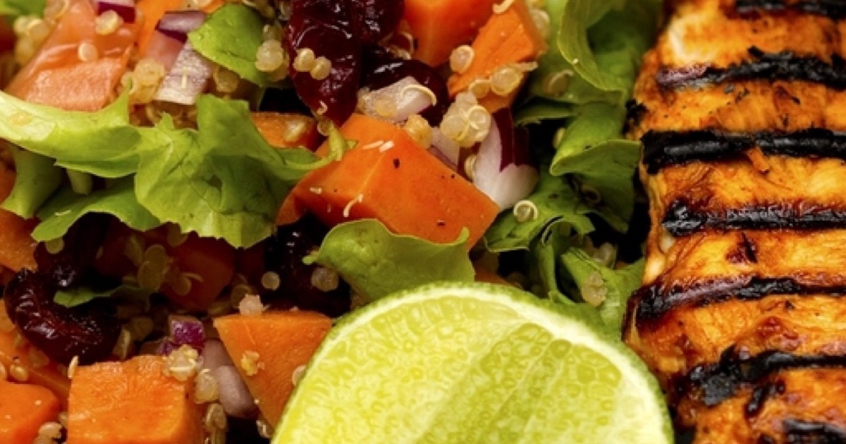 A close-up of grilled chicken breast served beside a quinoa salad with carrots, greens and a lime wedge.