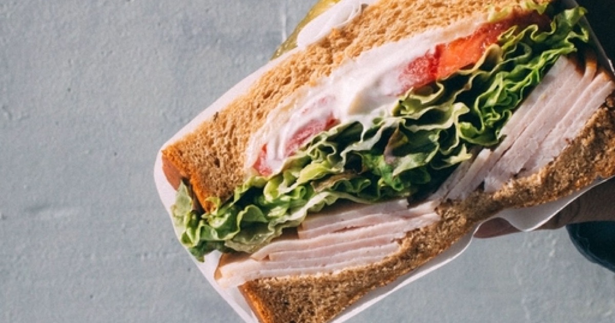 A hand holding a turkey, lettuce and tomato sandwich on wheat bread over a pale grey background.